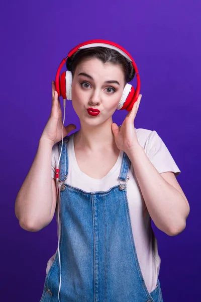Härlig flicka lyssna på musik i stora röda hörlurar isolerad på blå bakgrund på studio. — Stockfoto