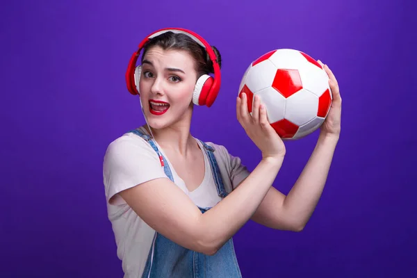 面白いです女の子歌で大赤いですヘッドフォンホールディングAサッカーボールで隔離された青の背景にスタジオ. — ストック写真