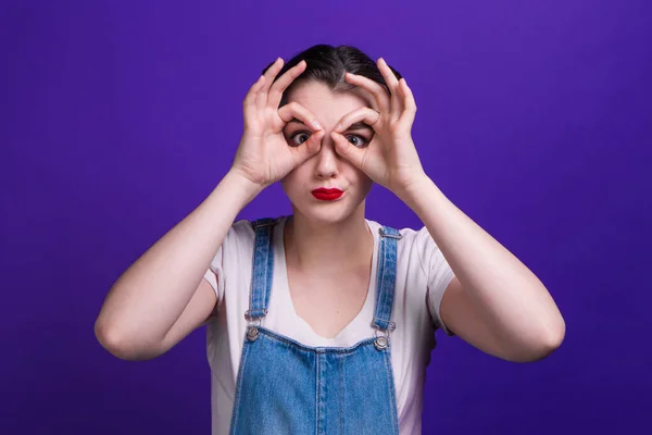 Bonita modelo olhando para a câmera através de seus dedos em gesto ok. Imitando binóculos isolados sobre fundo roxo . — Fotografia de Stock