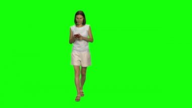 Genç kadın sakince yürüyor ve mesajlaşıyor. Cep telefonunu yeşil ekrana ver. Ön görünüm.