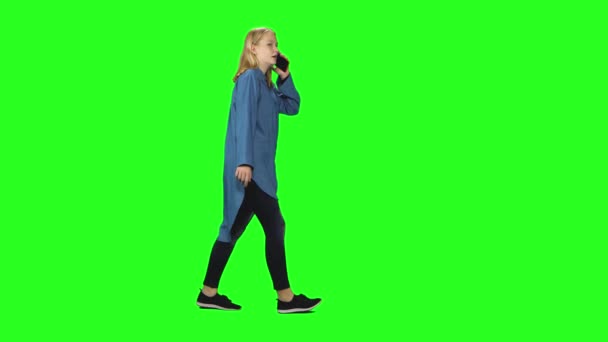 ブロンドのティーンエイジャーの女の子が落ち着いて歩いて、緑の画面上の携帯電話で話しています。クロマキー、 4kショット。プロフィールビュー. — ストック動画