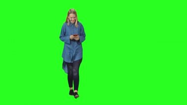 Sarışın genç kız yeşil ekranda sakince yürüyor ve cep telefonundan mesaj okuyor. Ön görünüm.