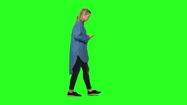 Chica adolescente rubia tranquilamente caminando y leyendo mensajes de texto en su teléfono móvil en la pantalla verde. Vista del perfil . — Vídeos de Stock