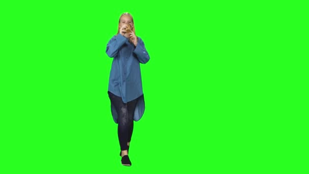 ブロンドのティーンエイジャーの女の子は、緑の画面の背景に落ち着いて歩いてコーヒーを飲んでいます。クロマキー、 4kショット。正面図. — ストック動画
