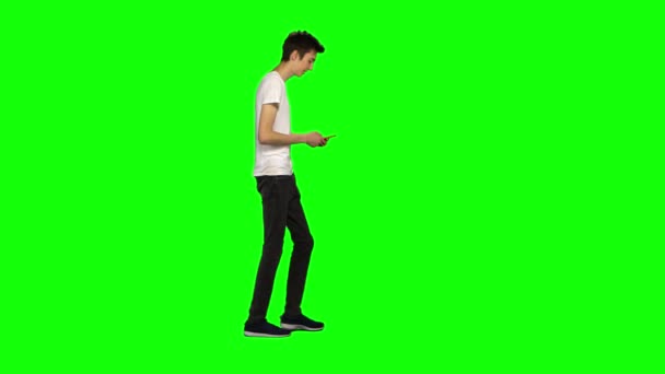 Tall skinny teen guy calmly walking and texting message vie his mobile phone on green screen. Chroma key. Profile view. — Stock Video