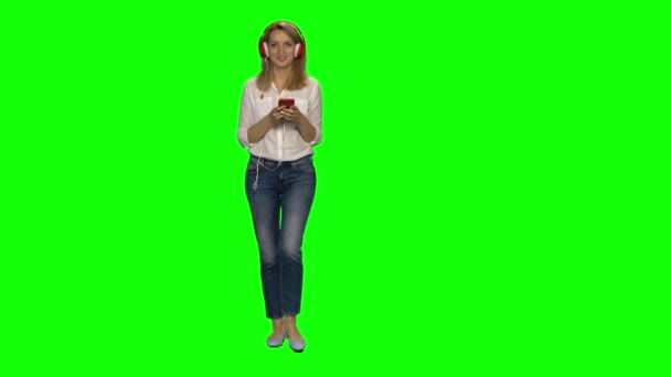 Blond kvinna går lugnt i stora röda hörlurar, väljer musik på mobilen på grön skärm. Chroma nyckel, 4k skott. Framifrån. — Stockvideo