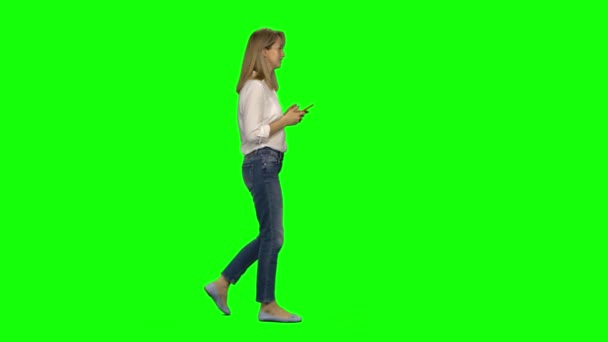 Blondine vrouw rustig wandelen en sms 'en met haar mobiele telefoon op greenscreen. Chroma sleutel, 4K schot. Profielweergave. — Stockvideo