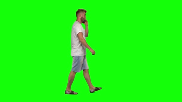 Бородатый парень спокойно ходит и разговаривает по мобильному телефону на зеленом фоне экрана. Chroma key, 4k shot. Профиль — стоковое видео