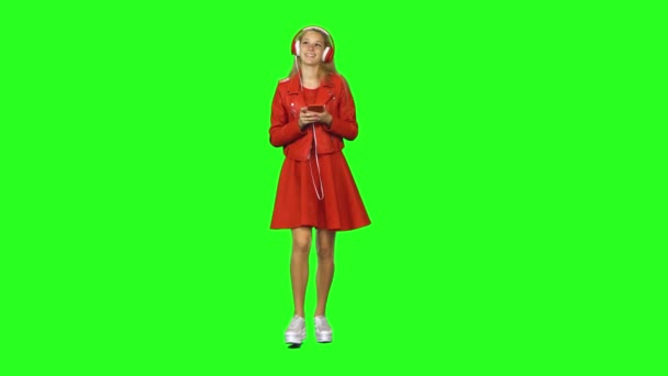 Blond meisje loopt in grote rode koptelefoon, kiest muziek en beweegt naar ritme op groen scherm — Stockvideo