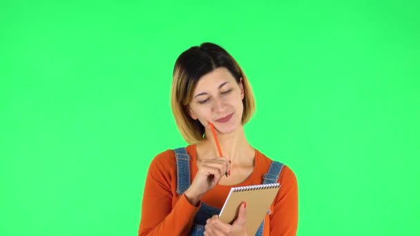Girl stands and thinks, then happy writes with pencil in notebook. Green screen — Stock Video