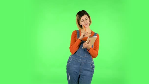 Girl stands and thinks, then happy writes with pencil in notebook. Green screen — Stock Video