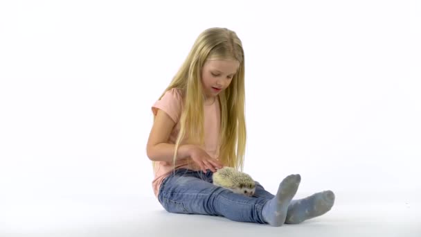 Маленькая девочка с длинными светлыми волосами держит ёжика на белом фоне. Медленное движение — стоковое видео