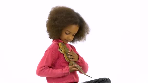 Malá africká kudrnatá holka drží a hladí vousatou agamu na bílém pozadí. Zpomalený pohyb — Stock video