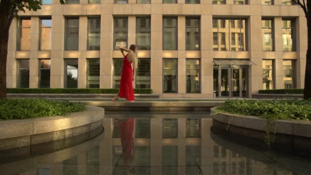 Красивая молодая девушка танцует на улице современного города и отражается в воде. На ней красное платье. Медленное движение. — стоковое видео