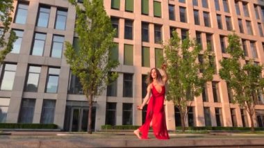 Günbatımında modern bir iş merkezi binasının caddesinde dans eden güzel bir genç kız. O kırmızı bir elbise giyiyor. Yavaş çekim.