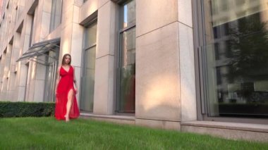 Modern bir şehrin arka planında yeşil çimlerde dans eden güzel bir genç kız. O kırmızı bir elbise giyiyor. Yavaş çekim.