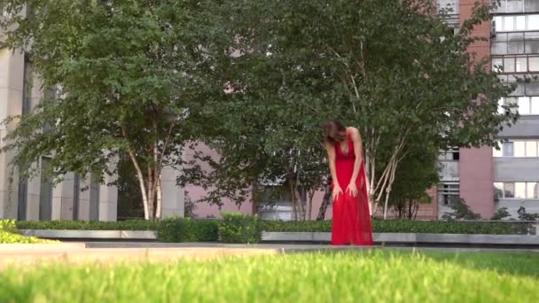 Όμορφη νεαρή κοπέλα χορεύει στο πράσινο γρασίδι στο παρασκήνιο μιας σύγχρονης πόλης. Φοράει ένα κόκκινο φόρεμα. Αργή κίνηση. — Αρχείο Βίντεο