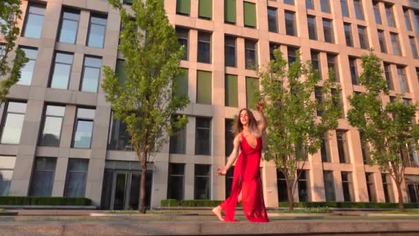 Beautiful young girl dancing on the street of a modern building of a business center in the sunset light. She is wearing a red dress. Slow motion. — Stock Video