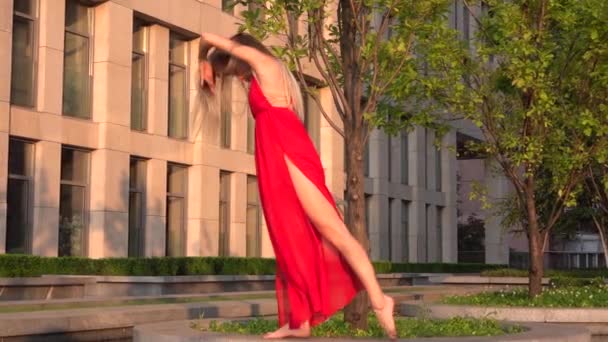 Hermosa joven bailando en la calle de un edificio moderno de un centro de negocios en la luz del atardecer. Lleva un vestido rojo. Movimiento lento . — Vídeos de Stock