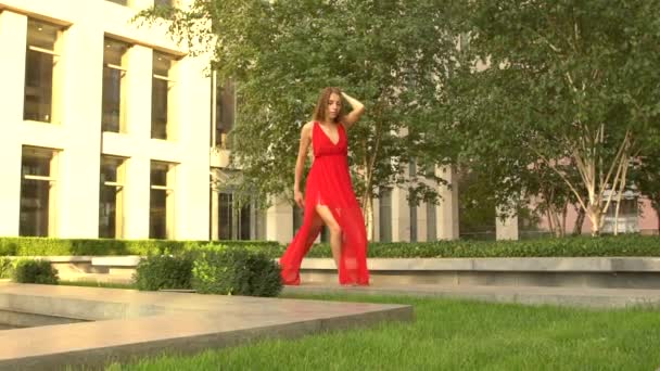 Vacker ung flicka dansar på grönt gräs mot bakgrund av en modern stad. Hon bär en röd klänning. Långsamma rörelser. — Stockvideo