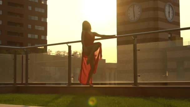 Prachtig jong meisje dansend op de straat van een moderne stad in het zonsondergang licht. Ze draagt een rode jurk. Langzame beweging. — Stockvideo