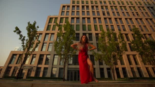 Красива молода дівчина танцює на вулиці сучасної будівлі бізнес-центру під час заходу сонця. Вона в червоній сукні. Повільний рух . — стокове відео