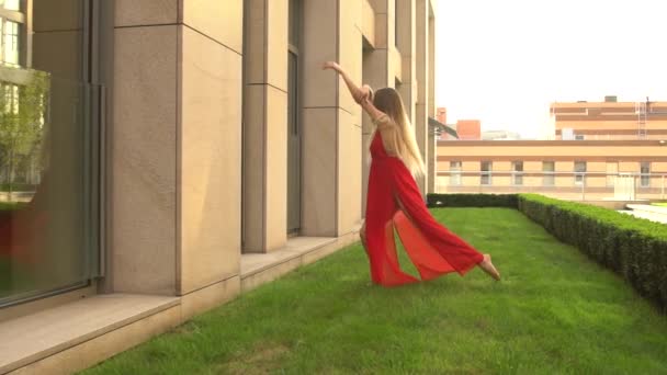 美しい若い女の子は、近代的な都市の背景に緑の芝生の上で踊る。彼女は赤いドレスを着ている。スローモーション. — ストック動画