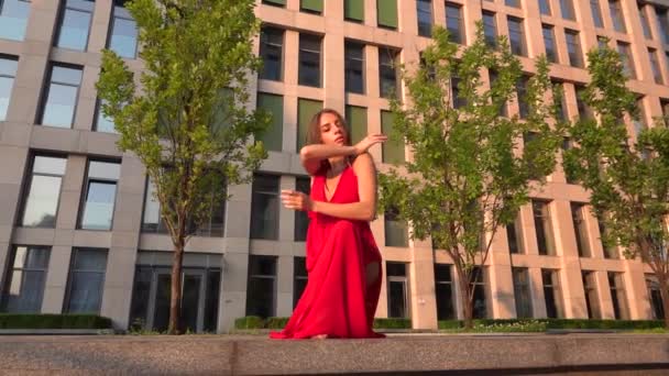 Menina bonita dançando na rua de um edifício moderno de um centro de negócios na luz do pôr do sol. Ela está usando um vestido vermelho. Movimento lento . — Vídeo de Stock