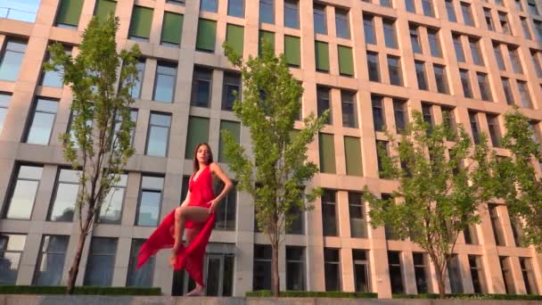 Beautiful young girl dancing on the street of a modern building of a business center in the sunset light. She is wearing a red dress. Slow motion. — Stock Video