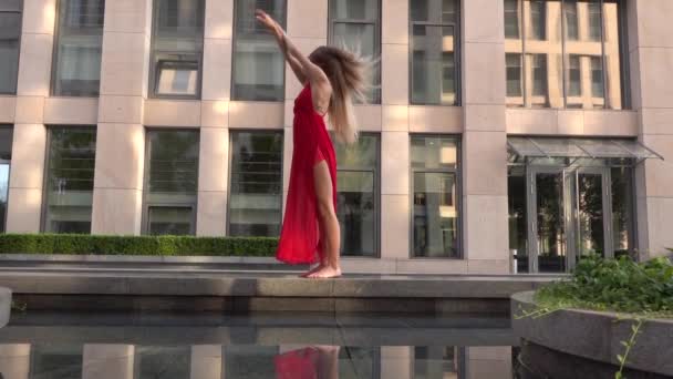 Beautiful young girl dancing on the street of a modern city and is reflected in the water. She is wearing a red dress. Slow motion. — Stock Video