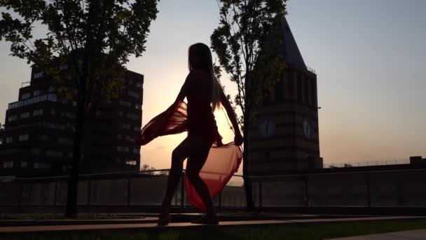 シルエット美しい若い女の子が夕日の光の中で近代的な都市の通りで踊ります。彼女は赤いドレスを着ている。スローモーション. — ストック動画