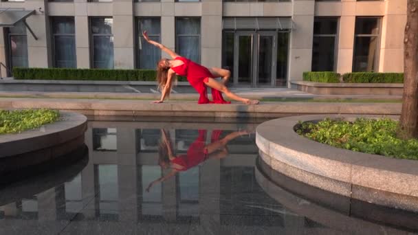 Hermosa joven bailando en la calle de una ciudad moderna y se refleja en el agua. Lleva un vestido rojo. Movimiento lento. — Vídeo de stock