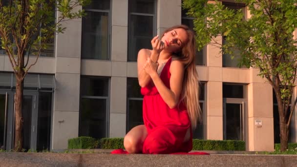 Hermosa joven bailando en la calle de un edificio moderno de un centro de negocios en la luz del atardecer. Lleva un vestido rojo. Movimiento lento . — Vídeo de stock