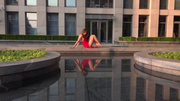 Hermosa joven bailando en la calle de una ciudad moderna y se refleja en el agua. Lleva un vestido rojo. Movimiento lento. — Vídeos de Stock