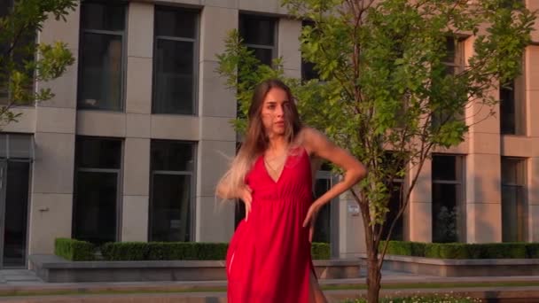 Hermosa joven bailando en la calle de un edificio moderno de un centro de negocios en la luz del atardecer. Lleva un vestido rojo. Movimiento lento . — Vídeos de Stock