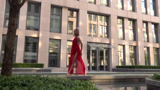 Prachtig jong meisje dansend op de straat van een moderne stad en wordt weerspiegeld in het water. Ze draagt een rode jurk. Langzame beweging. — Stockvideo