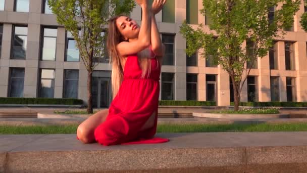 Beautiful young girl dancing on the street of a modern building of a business center in the sunset light. She is wearing a red dress. Slow motion. — Stock Video