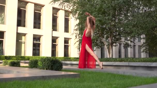 Vacker ung flicka dansar på grönt gräs mot bakgrund av en modern stad. Hon bär en röd klänning. Långsamma rörelser. — Stockvideo