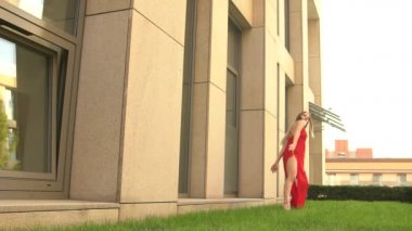 Modern bir şehrin arka planında yeşil çimlerde dans eden güzel bir genç kız. O kırmızı bir elbise giyiyor. Yavaş çekim.