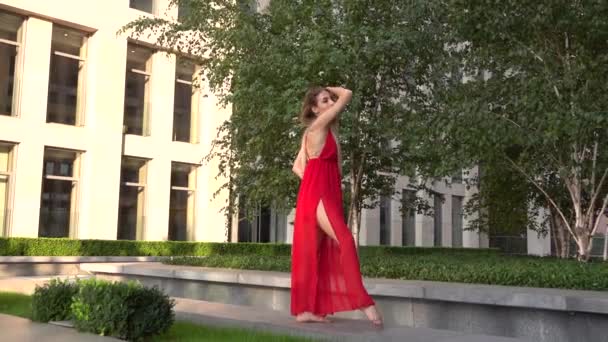 Menina bonita dançando na grama verde contra o fundo de uma cidade moderna. Ela está usando um vestido vermelho. Movimento lento . — Vídeo de Stock