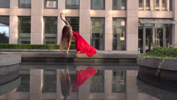 Beautiful young girl dancing on the street of a modern city and is reflected in the water. She is wearing a red dress. Slow motion. — Stock Video
