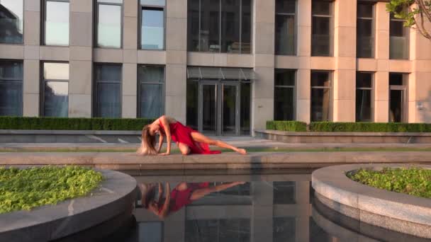 Belle jeune fille dansant dans la rue d'une ville moderne et se reflète dans l'eau. Elle porte une robe rouge. Mouvement lent. — Video