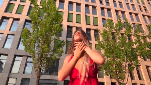 Beautiful young girl dancing on the street of a modern building of a business center in the sunset light. She is wearing a red dress. Slow motion. — Stock Video