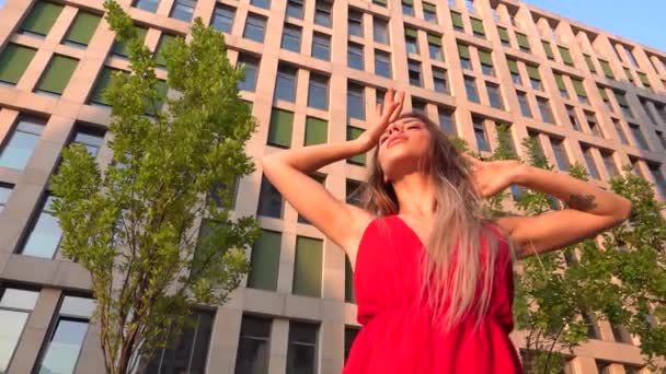 Beautiful young girl dancing on the street of a modern building of a business center in the sunset light. She is wearing a red dress. Slow motion. — Stock Video