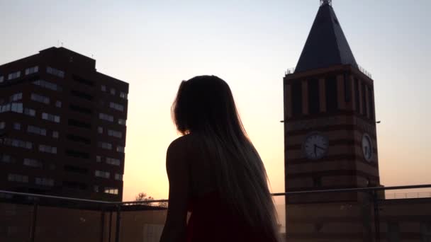 Σιλουέτα Όμορφη νεαρή κοπέλα χορεύει στο δρόμο μιας σύγχρονης πόλης στο φως του ηλιοβασιλέματος. Φοράει ένα κόκκινο φόρεμα. Αργή κίνηση. — Αρχείο Βίντεο