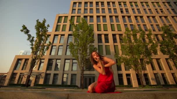 Hermosa joven bailando en la calle de un edificio moderno de un centro de negocios en la luz del atardecer. Lleva un vestido rojo. Movimiento lento . — Vídeos de Stock