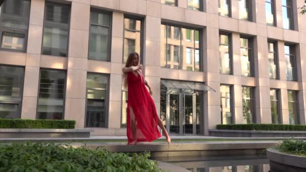 Krásná mladá dívka tančí na ulici moderního města a odráží se ve vodě. Má na sobě červené šaty. Zpomalený pohyb. — Stock video
