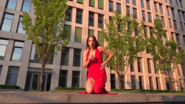 Menina bonita dançando na rua de um edifício moderno de um centro de negócios na luz do pôr do sol. Ela está usando um vestido vermelho. Movimento lento . — Vídeo de Stock