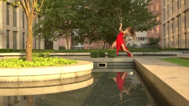 Hermosa joven bailando en la calle de una ciudad moderna y se refleja en el agua. Lleva un vestido rojo. Movimiento lento. — Vídeo de stock