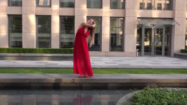 Beautiful young girl dancing on the street of a modern city and is reflected in the water. She is wearing a red dress. Slow motion. — Stock Video